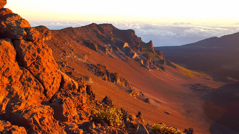 Golden hour lighting across the red volcanic landscape of Haleakalā Crater, viewed from the summit.