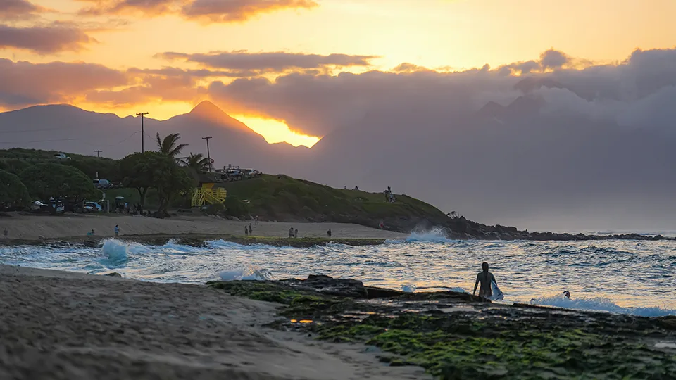 Sunset over Ho‘okipa Beach with a surfer silhouetted against the rocky shoreline.