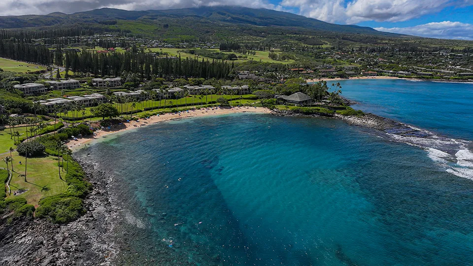 Aerial view of Kapalua Bay’s crescent-shaped shoreline framed by lush greenery and calm blue water.