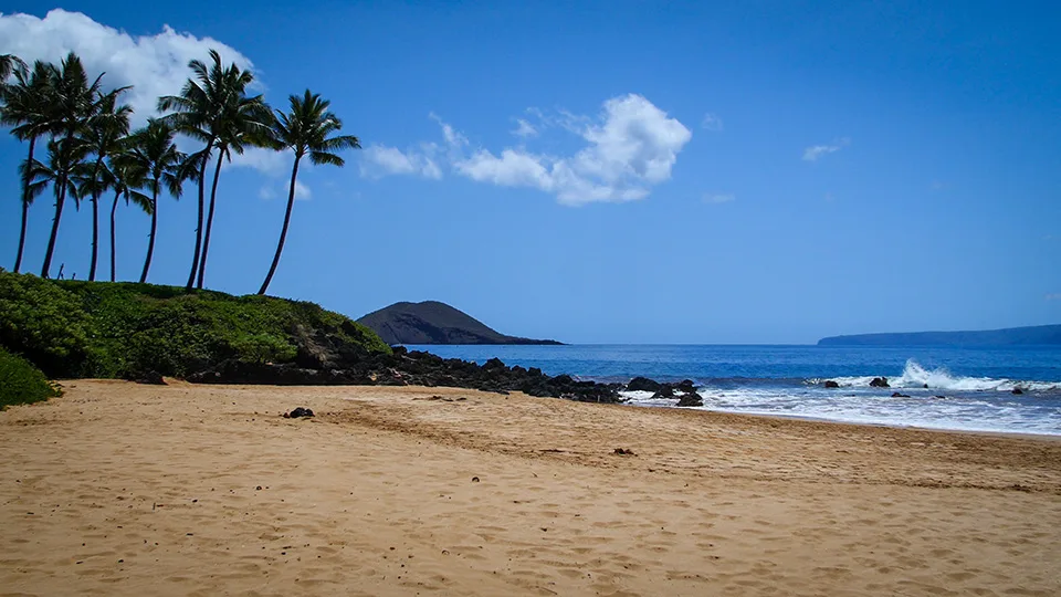 Poʻolenalena Beach on Maui with golden sand, palm trees, and Molokini Crater in the distance on a sunny day.